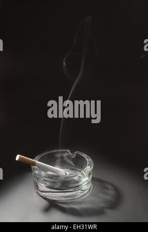 A lid cigarette in a glass ashtray with the smoke from the cigarette floating up in to the air. Stock Photo