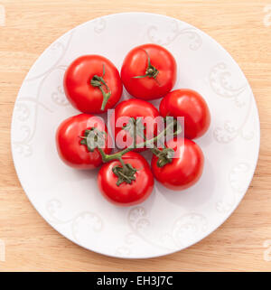 Large beefsteak tomatoes on the vine on a white plate, which is standing on a pale wood background. Stock Photo