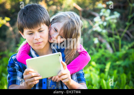 Teen boy with his younger sister sitting in a Park and using the tablet. Stock Photo