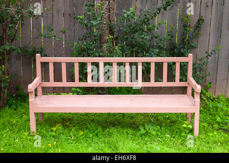 Old red bench stands on green grass near wooden fence in park Stock Photo