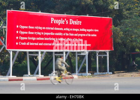 Man cycling in front of Tatmadaw (Army) Propaganda Poster titled 'People's Desire', Mandalay Stock Photo