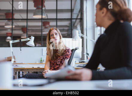 businesswoman speaking with her colleague in office. Young woman sitting at her desk discussing work with coworker. Stock Photo