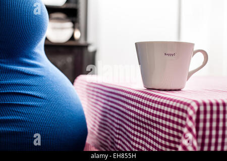 Woman in full term pregnancy wearing a blue top. The cup on the table reads happy.