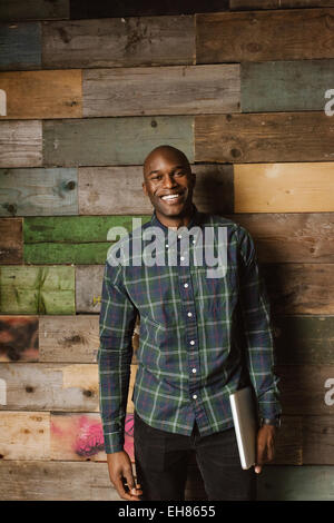 Portrait of happy young man holding a laptop looking at camera smiling while standing in office against a wooden wall. African a Stock Photo