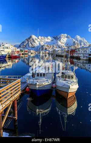 Boats docked in the calm waters of the port of Henningsvaer with the Norwegian Alps in the background, Lofoten Islands, Norway Stock Photo