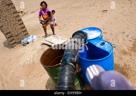 A Peruvian water distribution worker with a hose fills plastic barrels with drinking water on the dusty hillside in Lima, Peru. Stock Photo