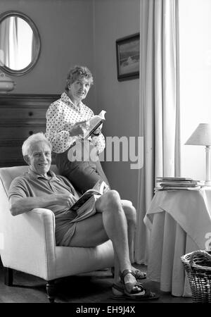 Retirement age couple in a front room positioned to a corner by the side of a window. The man is seated in a comfortable armchair, the woman standing and resting on the back of the chair by his side. The couple are holding books and looking into camera. They are relaxed and smiling. The man is wearing shorts and sandals, the woman trousers and long sleeved patterned blouse. The room setting is traditional, with curtain, a painting and mirror on the wall, a cloth covered occasional table and a wicker basket (partly cropped out of the shot). The image is black and white and portrait format. Stock Photo