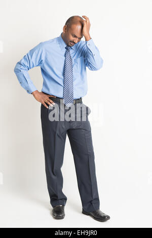 Full length Indian businessman hitting his head with hand, standing on plain background with shadow. Stock Photo