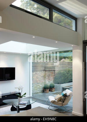 Living area with chair and flat TV screen with view through glass wall to outdoor courtyard in modern Walham Grove home, UK Stock Photo