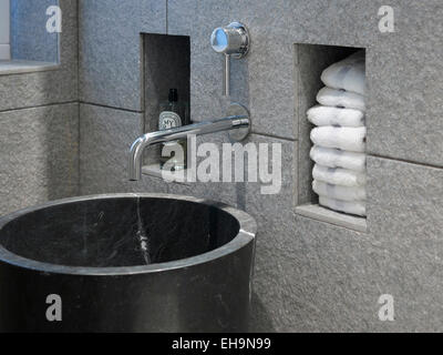 Close up of modern sculpted marble hand basin and running tap in stone tiled toilet with inset shelving for soap and towels in Cumberland Street home, UK Stock Photo