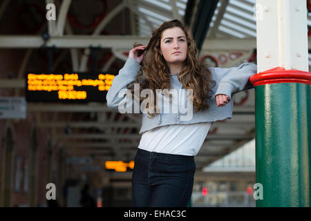 Leaving home: A sad looking lonely young 13 14 15 year old teenage girl with long brown hair outdoors  alone by herself  on a railway station platform waiting for a train  UK Stock Photo