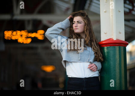 Leaving home: A sad looking lonely young 13 14 15 year old teenage girl with long brown hair outdoors  alone by herself  on a railway station platform waiting for a train  UK Stock Photo