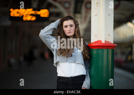 Leaving home: A sad looking lonely young 13 14 15 year old teenage girl with long brown hair outdoors  alone by herself  on a railway station platform waiting for a train  UK Stock Photo