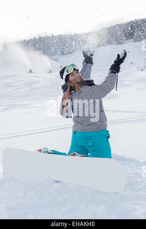 Young, curvy girl with snowboard, kneeling on the ski slopes throwing snow in the air. Stock Photo