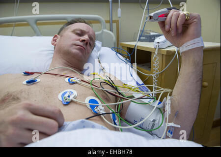 A man lies in a hospital bed with heart monitors to assess the risk of a heart attack, his pulse being taken on his finger Stock Photo