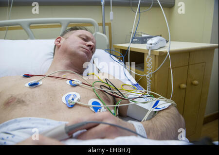 A man lies in a hospital bed with heart monitors to assess the risk of a heart attack, his pulse being taken on his finger Stock Photo