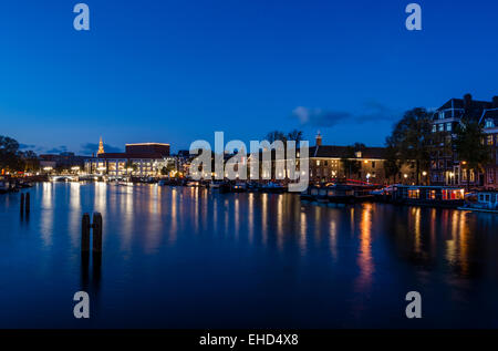 sA view of the river Amstel in Amsterdam after the sun has set and the sky is a deep blue. The city's skyline is illuminated. Stock Photo