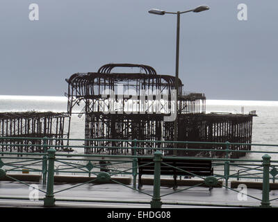 The Arson attacked ruin of the West Pier, Brighton designed by Eugenius Birch, seen across the promenade with decorative railings and benches to a glistening sea. Stock Photo
