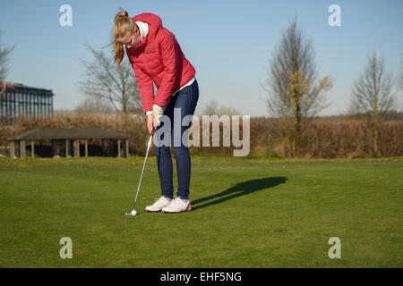 Woman playing golf lining up a putt on the green with her golf cart and clubs visible behind on a sunny blue sky day Stock Photo