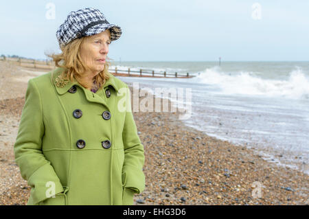 Elderly woman dressed warmly in the cold on a beach, looking out to sea. Stock Photo