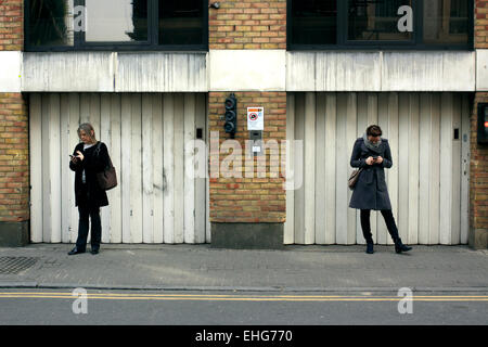 Two women checking their mobile phones on a street in Shoreditch, East London Stock Photo