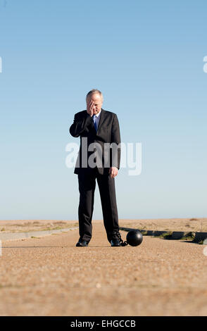 Businessman standing in the middle of a long road, head in hand and attached to a ball & chain. Stock Photo