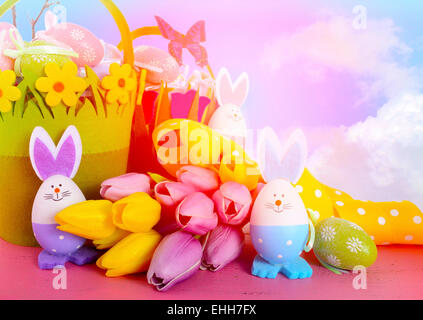 Happy Easter egg hunt baskets with tulip flowers and eggs with bunny ears and faces on a pink wood table, with sky background an Stock Photo