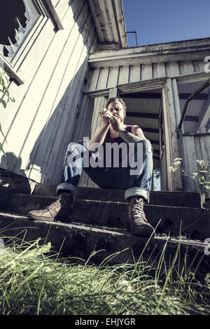 A young man sitting outside a abandoned house and lighten up a smoke. Stock Photo