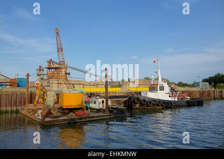 Working Tug moored alongside a cluttered jetty with a crane on a small barge. Stock Photo