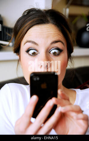 Young woman 20s looking shocked as she reads a message  on her Apple iPhone mobile phone at home Stock Photo