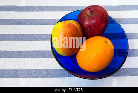 Two Apples and Orange in Blue Glass Bowl placed on Striped Background Stock Photo