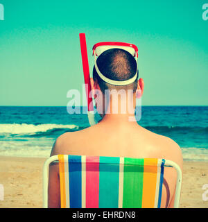 picture from the back of a man wearing a diving mask and a snorkel seated in a deckchair on the beach, with a retro effect