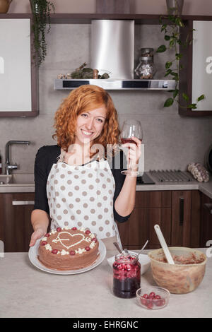 Happy redhead woman in love showing chocolate cake and holding glass of wine in the kitchen at home. Stock Photo