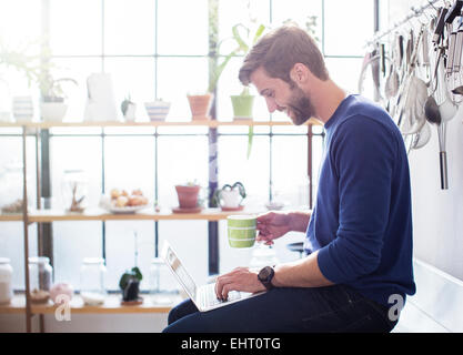 Young man sitting on kitchen counter with mug and laptop Stock Photo