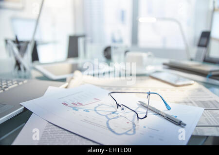 View of messy desk with glasses and pen on top of papers Stock Photo