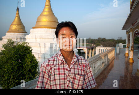 A young Burmese man with thanaka bark smeared on his face stands in front of golden spires of a Mandalay temple Myanmar Bagan Stock Photo