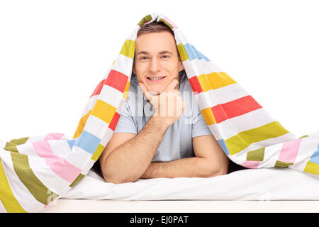 Studio shot of a carefree young man lying in a bed covered with striped multicolored blanket isolated on white background Stock Photo