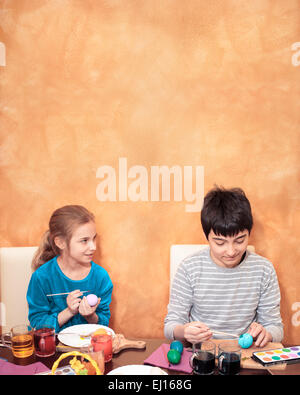 Boy and girl painting easter eggs at he table at home. Orange brushed wall as space for text in the background Stock Photo