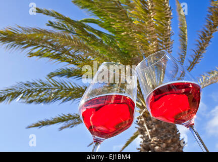 Two rosé wine glasses chinking 'cheers' in sunny holiday environment with palm tree and blue sky behind Stock Photo