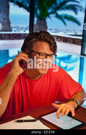 Happy mature man using his iPhone 6 smartphone and iPad air tablet computer in luxury vacation villa hotel interior pool at dusk Stock Photo