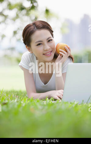 Young woman lying down on the lawn, holding an apple and using laptop with smile, Stock Photo