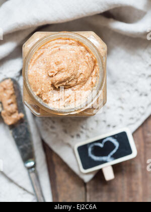 Vertical photo of homemade natural almond butter in a glass jar placed on rustic beige table cloth. View from above. Stock Photo