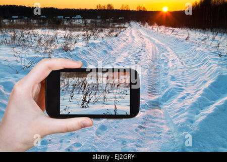 travel concept - tourist takes picture of sunset over blue winter snowdrifts on country field on smartphone, Stock Photo