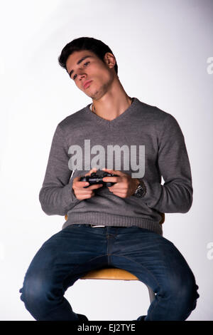 Attractive young man using joystick or joypad for videogames bored, on white background, sitting on stool Stock Photo
