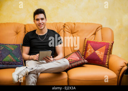 Handsome young man at home smiling, using tablet PC sitting on a couch Stock Photo