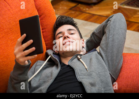 Handsome young man at home smiling, reading with ebook reader lying on a couch Stock Photo