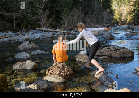 Nine year old girl balancing on her seven year old brother's shoulder as she climbs across rocks in the shallow river Stock Photo