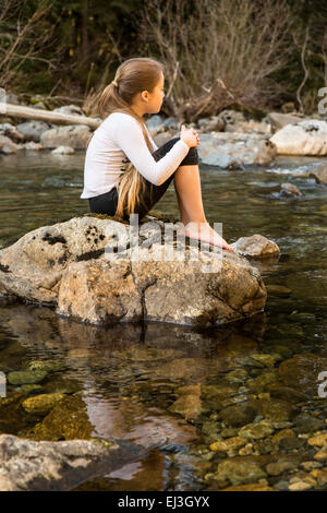 Nine year old girl daydreaming, sitting on a boulder in the shallow Snoqualmie river near North Bend, Washington, USA Stock Photo