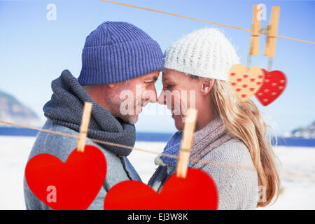 Composite image of attractive couple smiling at each other on the beach in warm clothing Stock Photo