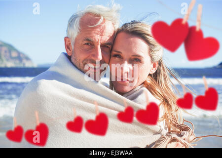 Composite image of smiling couple wrapped up in blanket on the beach Stock Photo